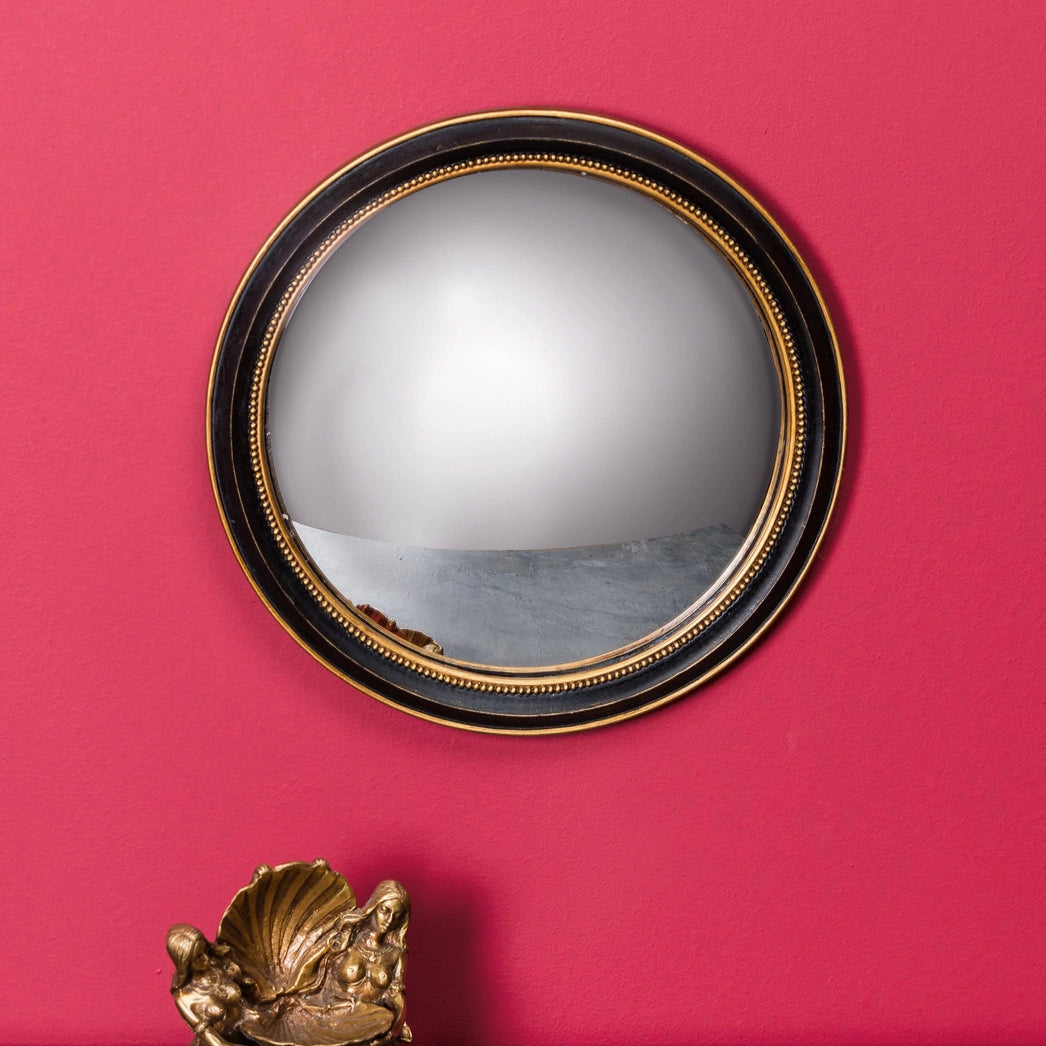 A black and gold convex mirror with a gold rim, placed against a red wall, with a decorative object to the bottom left.