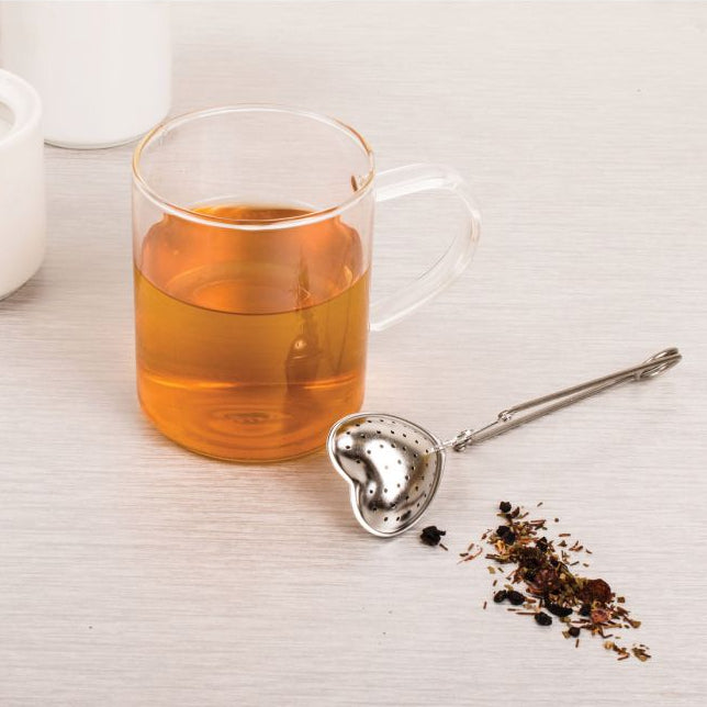 A clear glass mug filled with amber tea sits on a light wood surface, with a small metal heart-shaped tea infuser and scattered loose tea leaves beside it. A white container is partially visible in the background.