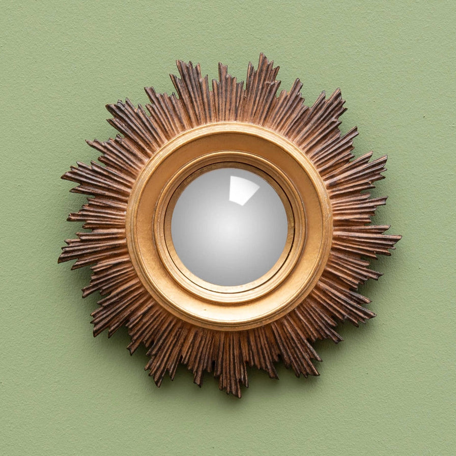 A round decorative wall mirror with a sunburst frame, featuring a warm gold-toned center and dark brown radiating spikes, mounted on a solid sage green wall.