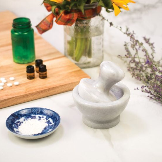 Mortar and pestle on a white surface with herbs and bottles in the background