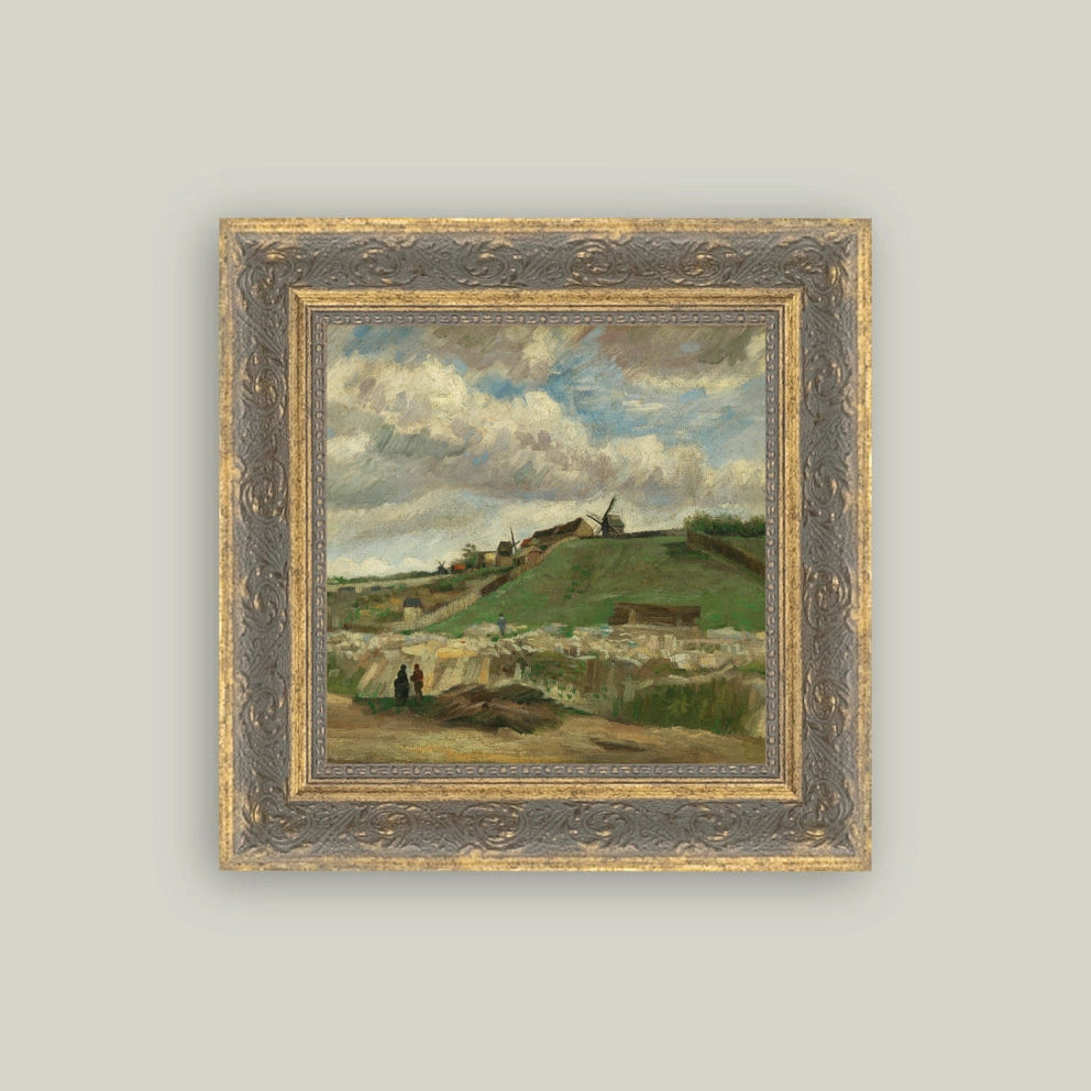A framed landscape painting featuring a green hillside village under a cloudy blue sky, set in an ornate gold and gray frame against a light gray background.