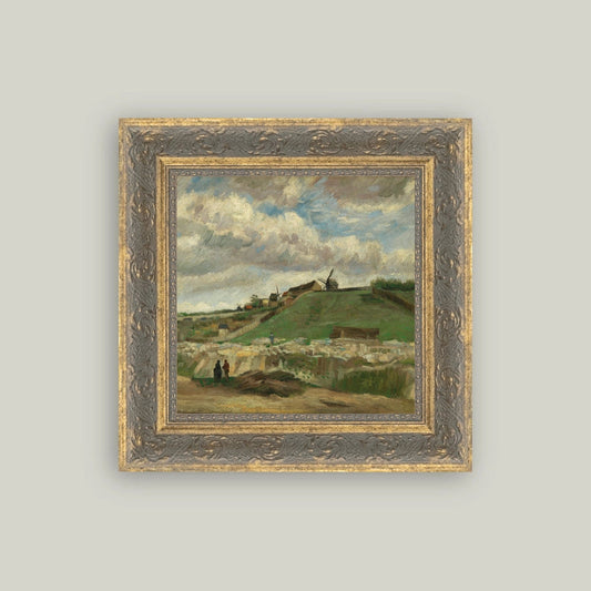 A framed landscape painting featuring a green hillside village under a cloudy blue sky, set in an ornate gold and gray frame against a light gray background.