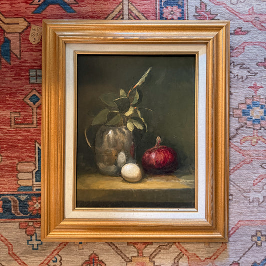 A framed still life oil painting featuring a glass vase with green leaves, a red onion, and an egg on a shelf, set in a gold frame against a patterned red and blue rug.