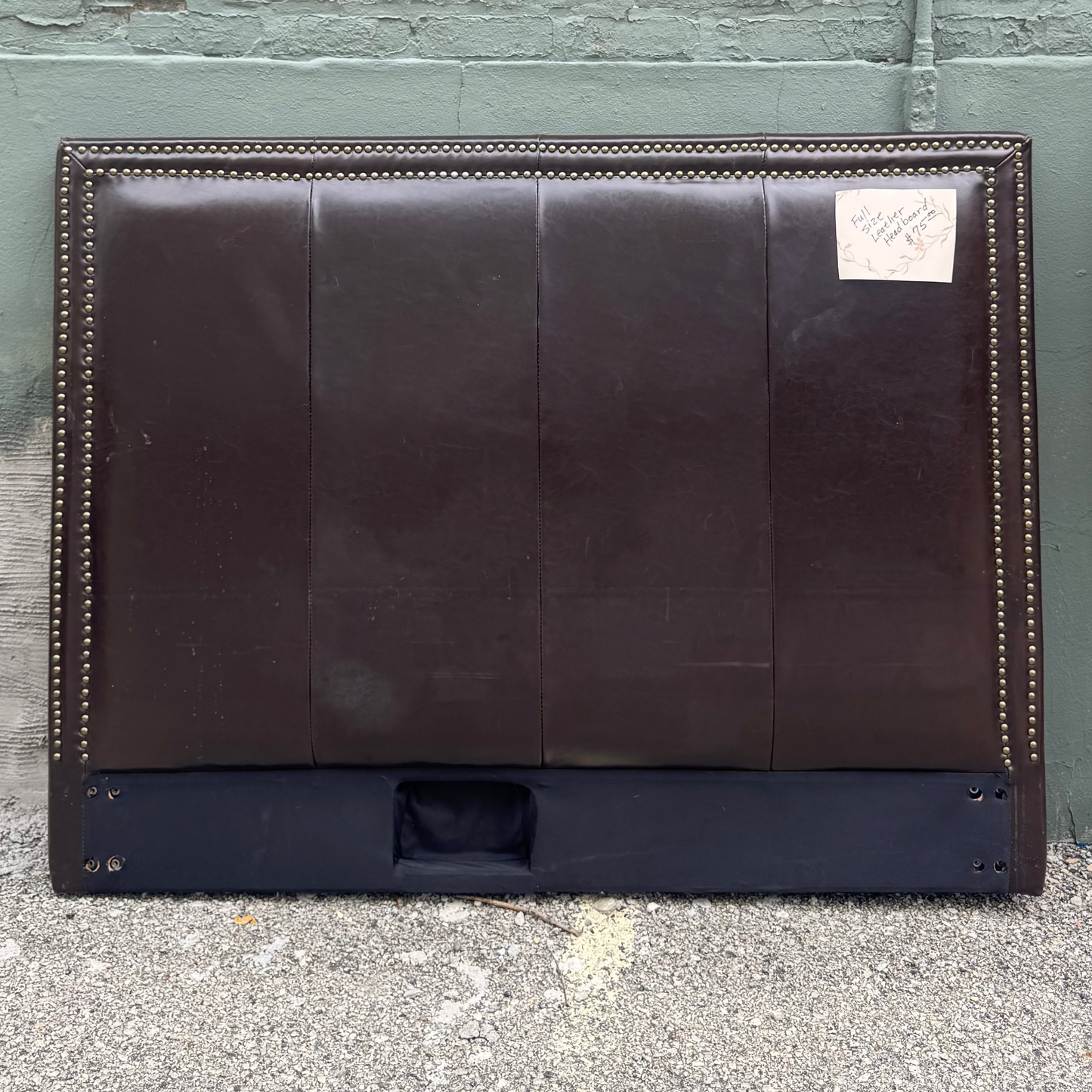 A dark brown leather headboard with button tufting and brass nailhead trim, leaning against a distressed green brick wall on a concrete floor.