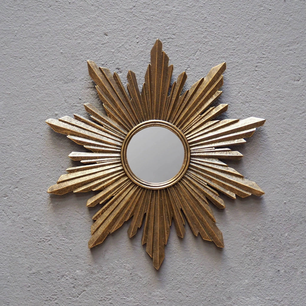 A gold sunburst wall mirror with a circular reflective center, mounted on a textured light gray plaster wall.