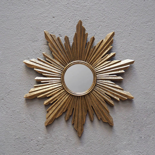 A gold sunburst wall mirror with a circular reflective center, mounted on a textured light gray plaster wall.
