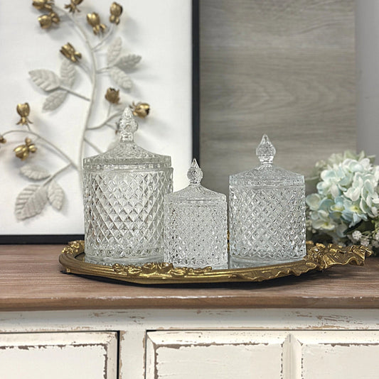 Decorative glass jars on a gold tray with a framed artwork and flowers in the background.